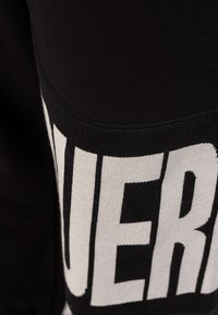 Gros plan sur un tissu noir avec un texte blanc audacieux. Matière douce et extensible avec une section de couleur contrastante, mettant en avant le design graphique.