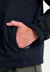 Schwarze Jacke mit glatter Textur, ausgestattet mit einem elastischen Bündchen und einer Hand in der Seitentasche. Es gibt ein kleines Reißverschlussdetail nahe dem Saum.