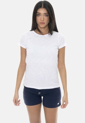 Femme aux longs cheveux foncés portant un t-shirt blanc uni et un short bleu marine, debout devant un fond blanc.
