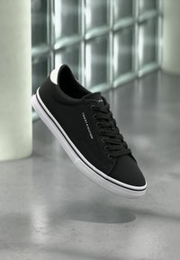 Μαύρο αθλητικό παπούτσι με λευκές λεπτομέρειες, φτιαγμένο από λείο ύφασμα. Διαθέτει στρογγυλές μύτες, κορδόνια στο μπροστινό μέρος και ορατό λογότυπο της μάρκας στο πλάι.