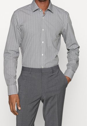 Man met een wit overhemd met lange mouwen en een zwart cirkelpatroon, en grijze pantalon, met één hand in de zak, voor een witte achtergrond.