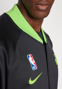 Fekete zipzáras dzseki világos zöld gallérral. Az NBA logója és a Nike pipa zöld színben van, így passzolnak egymáshoz. Az anyag simának és sportosnak tűnik.