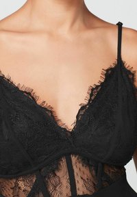 Bralette de encaje negro con escote pronunciado, con bordes festoneados, tirantes ajustables y un panel de encaje transparente en el centro.