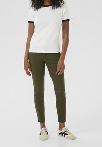 T-shirt blanc avec bordure noire, assorti à un pantalon ajusté vert olive et des baskets blanc cassé avec des accents noirs. Tenue simple et décontractée.