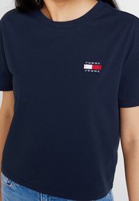 Námořnická modrá tričko Tommy Jeans na osobě, upravené v pase, spárované se světle modrými džíny.