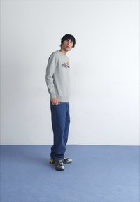 Grijze sweatshirt met een logo, gecombineerd met losse blauwe spijkerbroeken en zwarte sneakers. Staande pose tegen een lichte achtergrond.