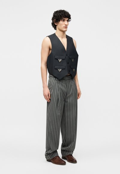 Homme portant un gilet noir sans manches avec quatre poches à l'avant, un pantalon large rayé noir et blanc, et des chaussures marron sur un fond uni.