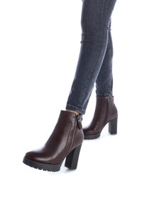 Botas de cuero marrón hasta el tobillo con un alto tacón cuadrado, cremallera lateral y detalles texturizados en el panel del tacón. Llevadas con jeans de mezclilla oscuros.