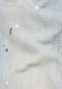 Tejido azul claro con una textura entrelazada; presenta manchas de pintura marrón y blanca dispersas por toda la superficie. Superficie suave y ligeramente arrugada.