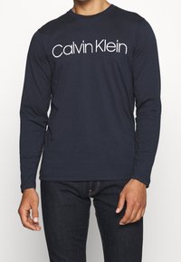 Granatowa koszulka z długim rękawem wykonana z bawełny. Posiada biały logo "Calvin Klein" na wysokości klatki piersiowej. Klasyczny fason z okrągłym dekoltem.