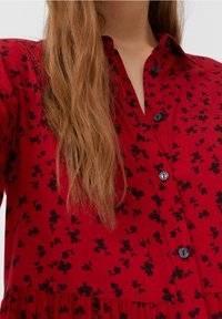 Robe rouge ornée d'un motif floral noir, avec un design à boutons sur le devant et un col. Le tissu semble lisse avec un léger éclat.