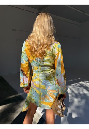 Kobieta o długich blond włosach, ubrana w kolorową sukienkę z tropikalnym wzorem, trzymająca teksturowaną plecioną torebkę, stojąca w nasłonecznionym pokoju.