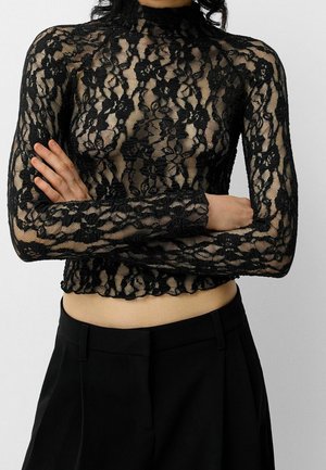 Frau trägt ein schwarzes, florales Spitzen-Crop-Top mit langen Ärmeln und hohem Kragen, kombiniert mit hoch taillierter schwarzer Hose, Arme verschränkt.