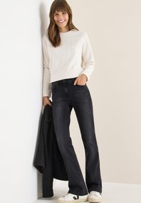Jasny sweatshirt z wytłoczonym tekstem, ciemne jeansy o prostym kroju i białe trampki. Model trzyma czarny płaszcz. Prosty, codzienny strój.
