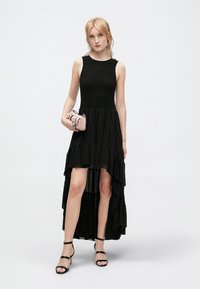 Schwarzes ärmelloses Kleid mit taillierter Passform, gestuftem Saum und asymmetrischem Schnitt. Glattes Material mit fließender Textur. Kombiniert mit einer rosa Handtasche.