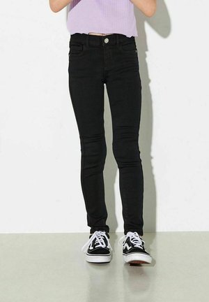 Person trägt schwarze Skinny-Jeans, schwarz-weiße Sneakers und ein bauchfreies lavendelfarbenes Rippentop, steht vor einer weißen Wand.