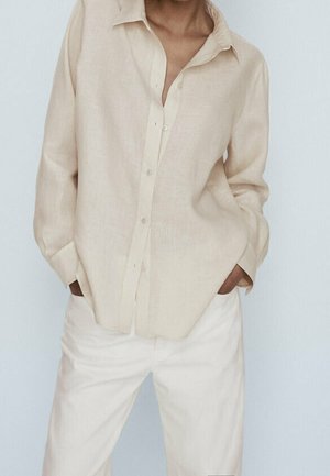 Person wearing a loose beige button-up shirt with sleeves rolled down and light-colored pants, hands in pockets against a plain background.