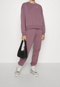 Lila sweatshirt med kort design och broderad logotyp, tillsammans med matchande joggers. En svart handväska och ljusa sneakers fulländar looken.