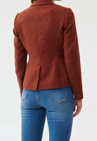 Blazer ajusté couleur rouille en tissu texturé, doté d'un col cranté, d'une coupe slim et d'une fente au dos. Associé à un jean bleu.