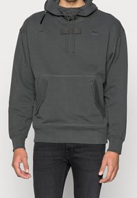 Felpa grigia con cappuccio regolabile con coulisse, tasche a marsupio frontali e polsini a coste. Presenta una patch con logo sulla parte superiore sinistra. Texture liscia.