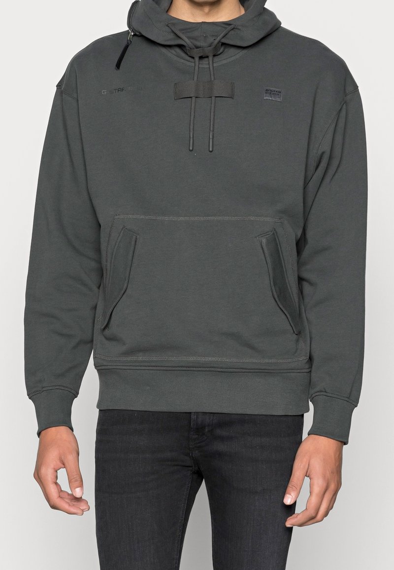 Felpa grigia con cappuccio regolabile con coulisse, tasche a marsupio frontali e polsini a coste. Presenta una patch con logo sulla parte superiore sinistra. Texture liscia.