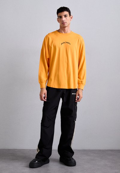 Tricou cu mânecă lungă, de culoare portocalie, cu text negru "palm Angels", asortat cu pantaloni cargo negri, largi, cu dungi pe laterale și buzunare.