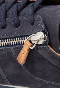 Sapata de camurça azul-marinho com um puxador de zipper em couro de contraste, zipper prateado e atacadores azul-marinho. Apresenta detalhes costurados ao longo das laterais.