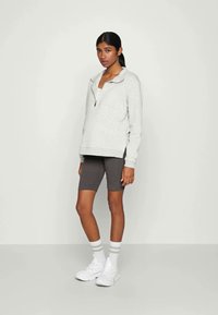 Felpa zip grigia chiara con tasca frontale, abbinata a pantaloni da bicicletta grigi scuri. Sneakers bianche con calzini a righe grigie e bianche.