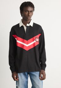 Giovane uomo che indossa una maglietta da rugby nera con colletto bianco e righe a zigzag rosse e bianche, abbinata a jeans azzurri sbiaditi.