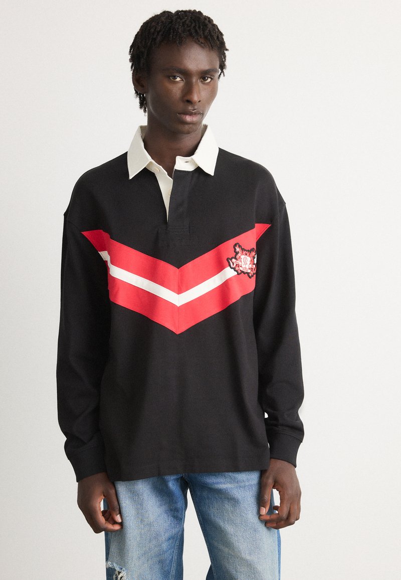 Giovane uomo che indossa una maglietta da rugby nera con colletto bianco e righe a zigzag rosse e bianche, abbinata a jeans azzurri sbiaditi.