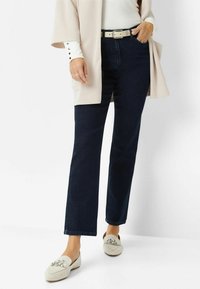 Jeans dritti blu scuro abbinati a una maglietta crema e un cardigan beige chiaro. Le scarpe bianche presentano un accento decorativo in argento.