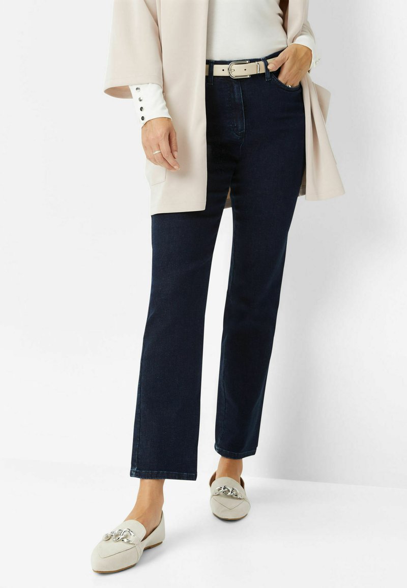Jeans dritti blu scuro abbinati a una maglietta crema e un cardigan beige chiaro. Le scarpe bianche presentano un accento decorativo in argento.