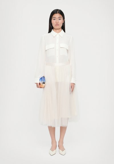 Blusa blanca transparente con dos bolsillos en el pecho, combinada con una suave falda de tul con capas. La modelo sostiene un clutch metálico y lleva tacones blancos de punta.