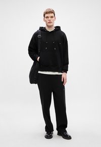Sudadera negra con cordones, combinada con pantalones negros holgados. El modelo lleva un bolso negro y calza zapatos negros. Diseño simple y mínimos detalles.