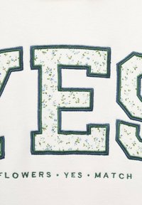 Bela tkanina z velikim napisom "DA" v cvetličnem vzorcu, ki vključuje majhne modre cvetlice in zelene liste, obšit z temno zelenim šivom.