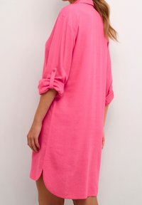 Różowa, sięgająca do kolan sukienka koszulowa z lekkiego materiału, z kołnierzykiem, rękawami z możliwością podwinięcia oraz bocznymi rozcięciami dla większej swobody ruchów.