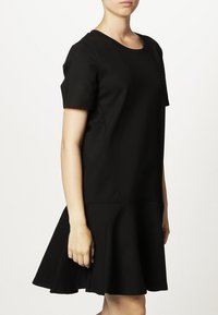 Femme portant une robe noire à manches courtes avec un ourlet évasé, se tenant devant un fond blanc uni.