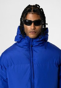 Blauwe pufferjack met hoge kraag, ritssluiting en zijzakken. Draag met zwarte zonnebril. Textuurstof, geïsoleerd.