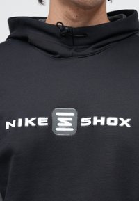 Μαύρη φούτερ με κουκούλα και κορδόνι, με μεγάλο λευκό λογότυπο "NIKE SHOX" στο στήθος. Το υλικό φαίνεται απαλό με υφή τελειώματος.
