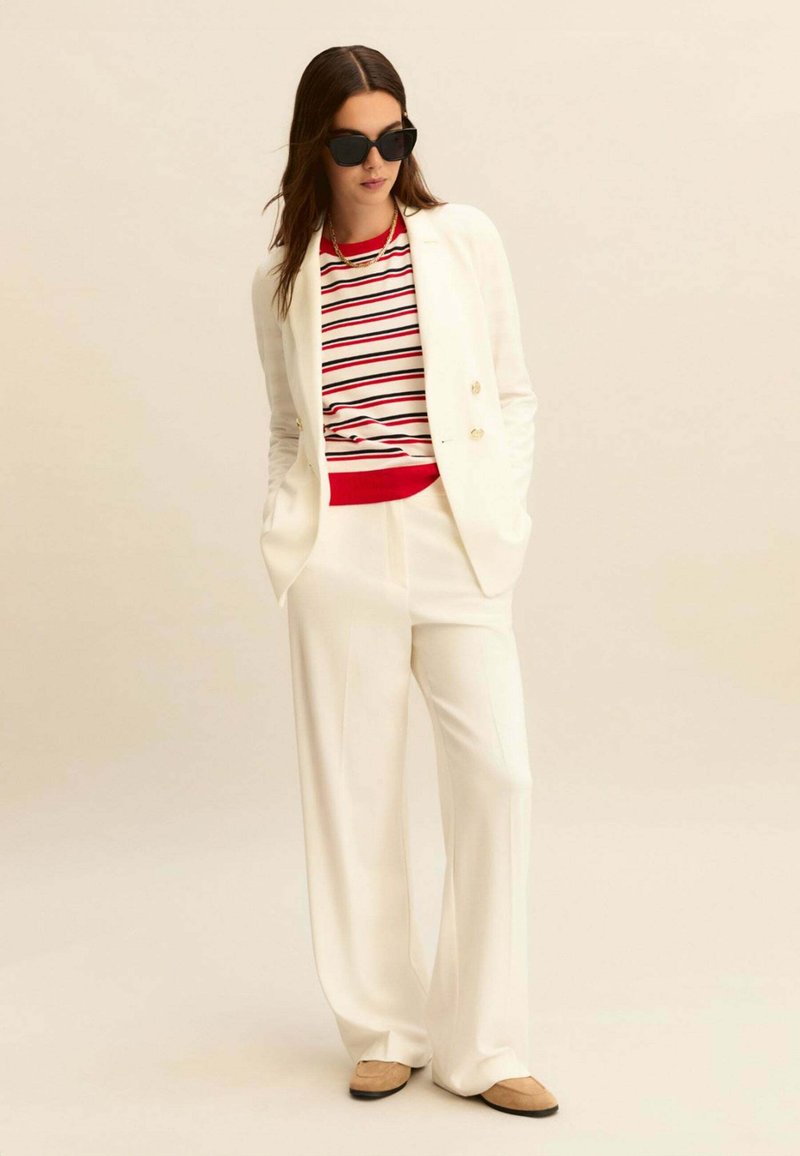 Donna con blazer bianco e pantaloni a gamba larga, maglione a righe rosse, bianche e nere, scarpe beige, occhiali da sole neri e collana d'oro.