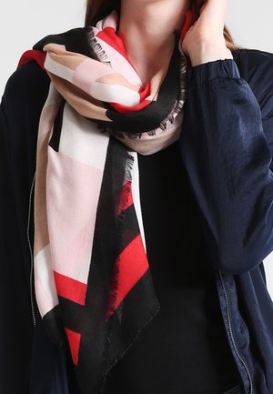 Personne portant une écharpe à motifs géométriques multicolores en rouge, noir, beige et rose, par-dessus un haut noir et une veste bleu marine.