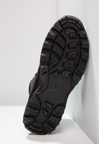 Crna Nike potplat s dubokim utorima koji se oslanja na bijeli zid i pod.