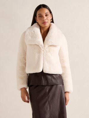 Witte nepbont jas met een brede kraag en lange mouwen, met een cropped ontwerp. Draagt over een donkerbruine, aansluitende leren rok.