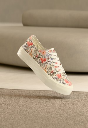 Low-top white sneaker with pink and orange floral print, white laces, and thick rubber sole placed on beige carpet in front of a sofa.