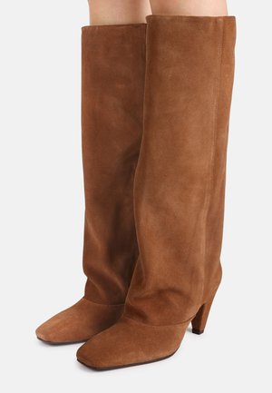 Bottes montantes en daim marron avec bout carré et talon bloc portées par une personne aux jambes nues sur un fond blanc.