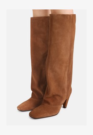 Botas altas marrones de ante con punta cuadrada y tacón grueso, usadas por una persona con las piernas descubiertas sobre un fondo blanco.