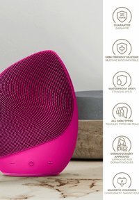 Dispositif de nettoyage facial en silicone rose avec une surface texturée, une forme arrondie et des commandes à l'avant. Il est étanche (IPX7) et dispose d'une charge magnétique.