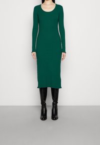 Långärmad, ribbad klänning i mörkgrön, knälång med en rundad halsringning och råa fållar, kombinerad med knähöga svarta stövlar.