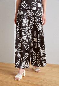 Czarne szerokie spodnie z białymi kwiatowymi wzorami, wykonane z gładkiej tkaniny. Połączone z białymi plecionymi sandałami na słupku.