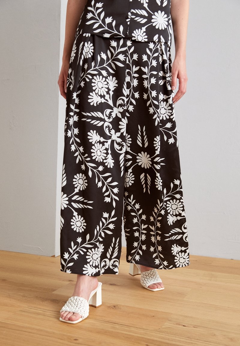 Czarne szerokie spodnie z białymi kwiatowymi wzorami, wykonane z gładkiej tkaniny. Połączone z białymi plecionymi sandałami na słupku.